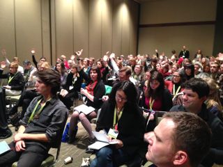 Audience members raise their hands in response to a presenter's question.